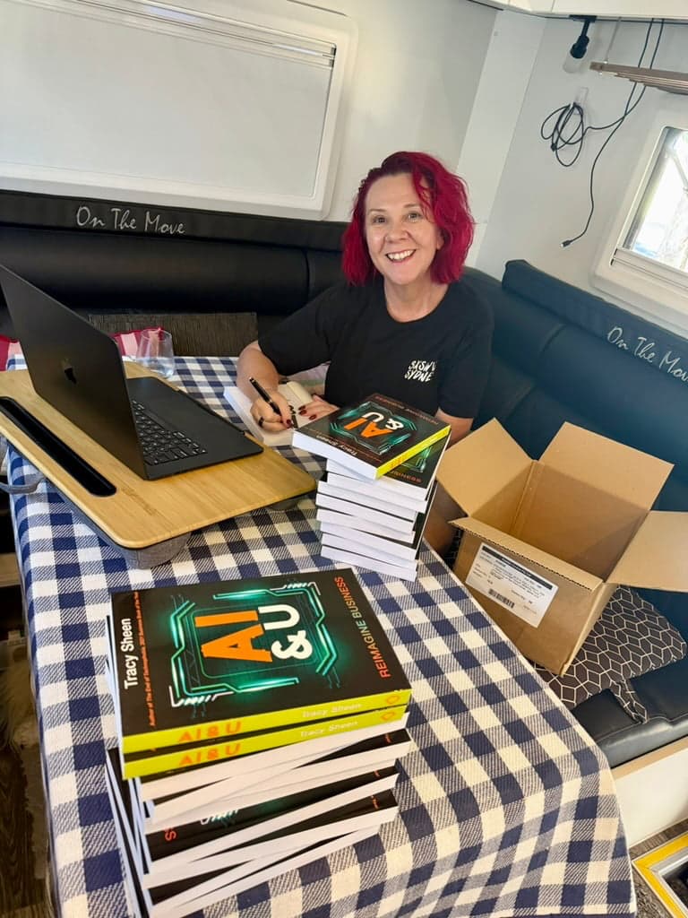 Tracy Sheen in her caravan signing copies of AI & U, with a stack of paperbacks ready to ship.