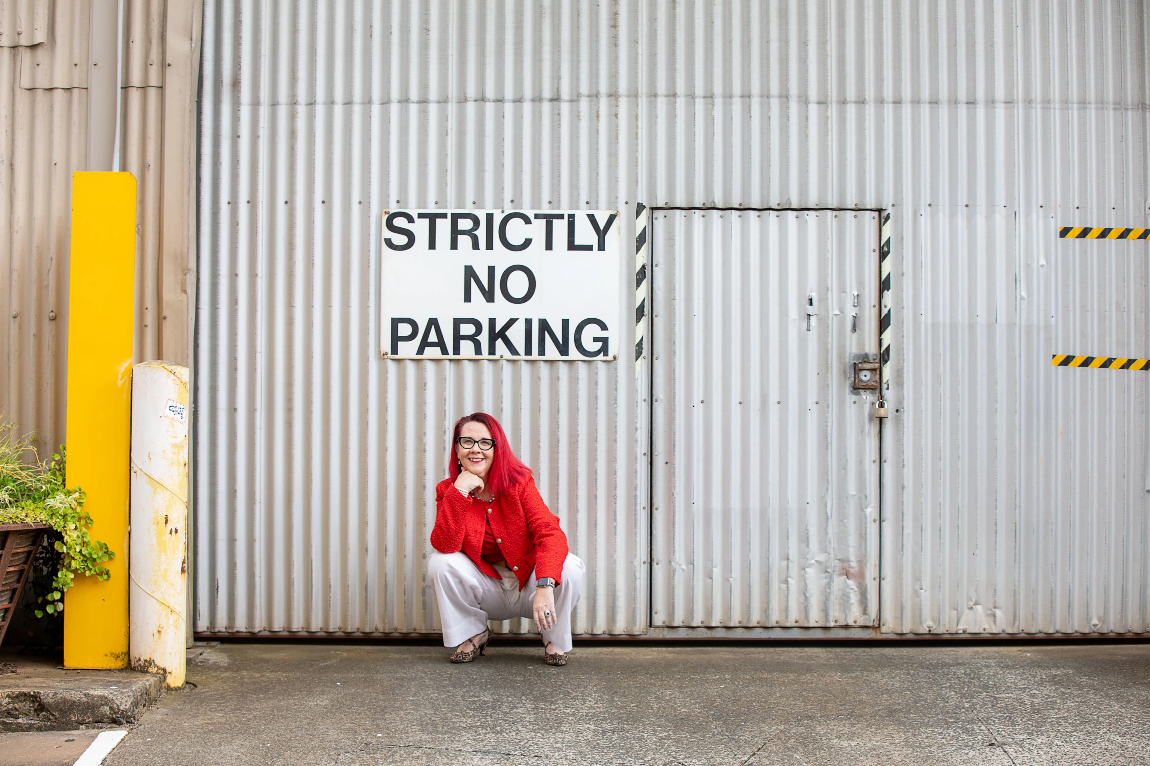 Tracy Sheen in a red blazer and white pants standing beside a Strictly No Parking sign on an industrial corrugated metal door.